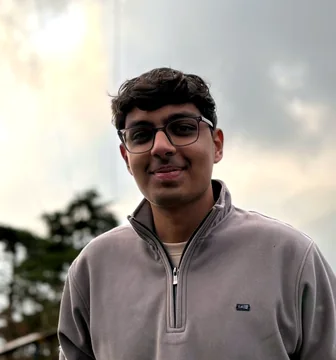 Portrait of Shreyansh Modi, smiling, wearing glasses and a grey zip-up sweater outdoors.